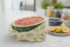 Half-cut watermelon wrapped in a reusable beeswax food wrap on a white surface with fruits in the background.